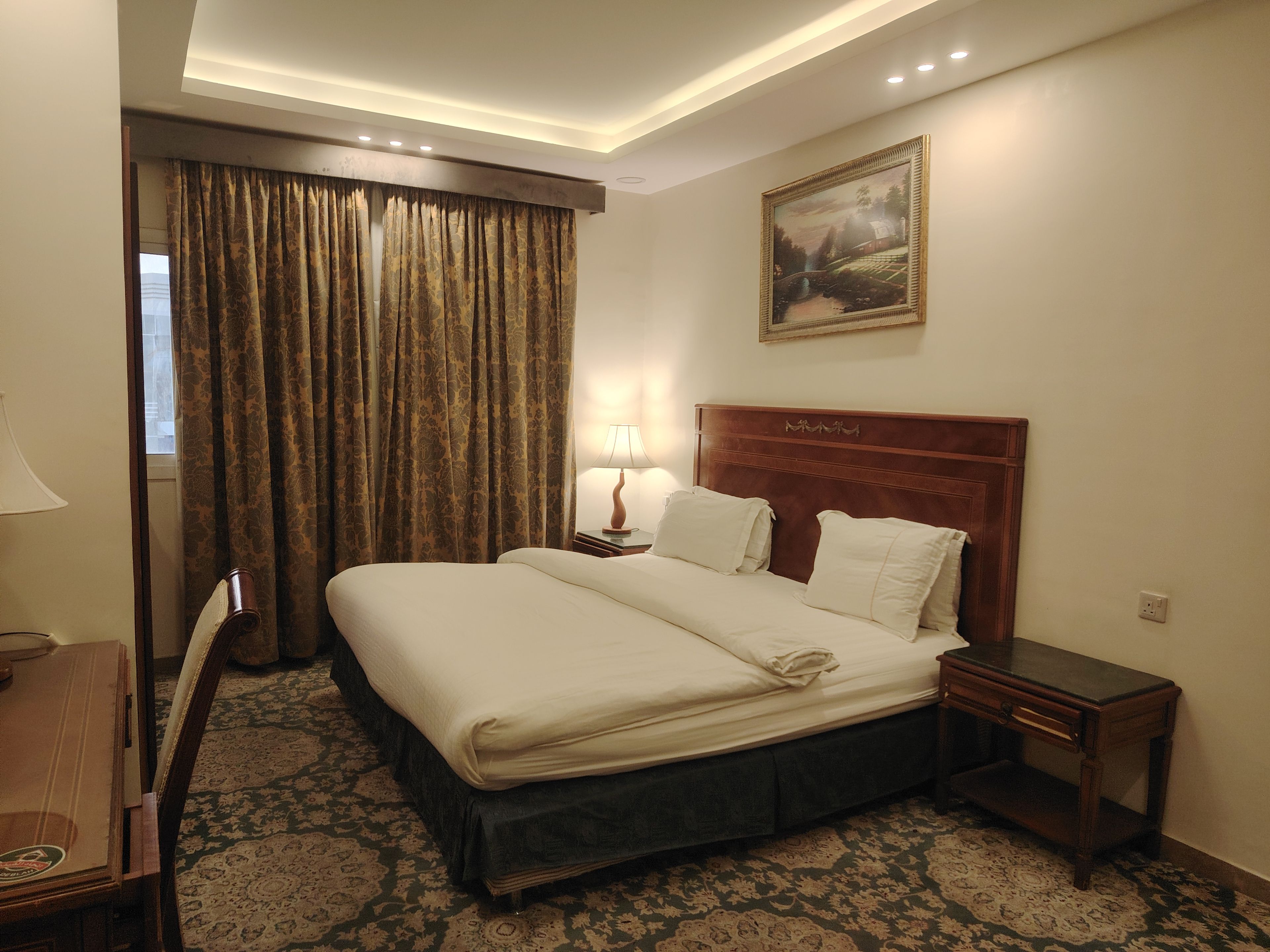 Una habitación de hotel con una cama grande, una mesita de noche, una lámpara, un cuadro en la pared y una alfombra estampada.