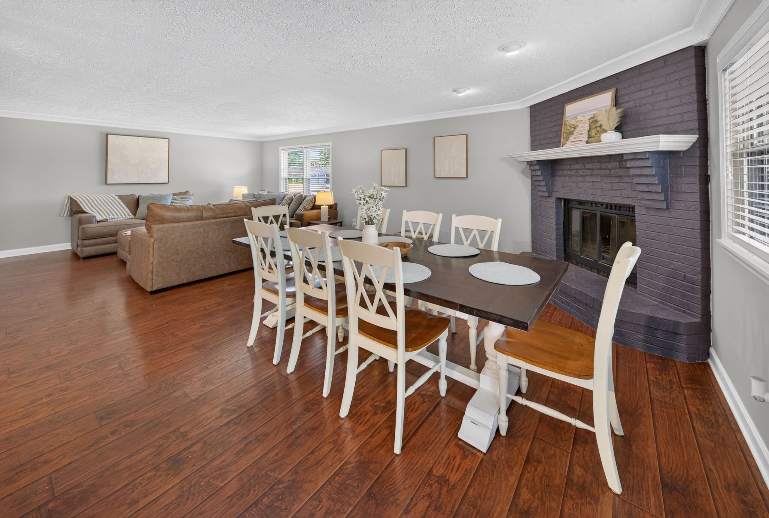Un comedor con mesa de madera y sillas blancas, chimenea y sala de estar con sofá y lámpara.