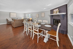 Un comedor con mesa de madera y sillas blancas, chimenea y sala de estar con sofá y lámpara.
