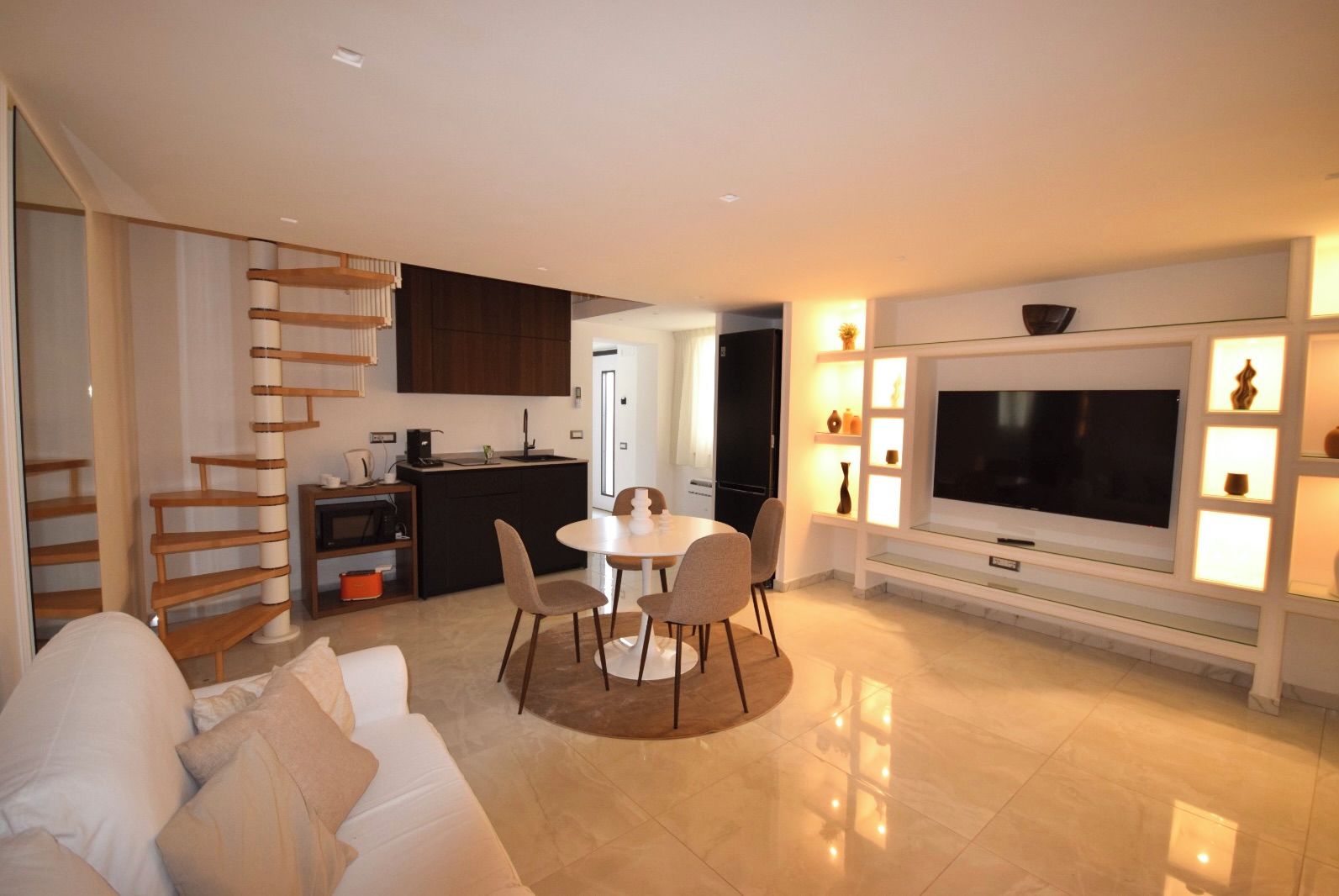 Un salon moderne comprenant un canapé, une table à manger avec des chaises, un coin cuisine et un téléviseur fixé au mur.