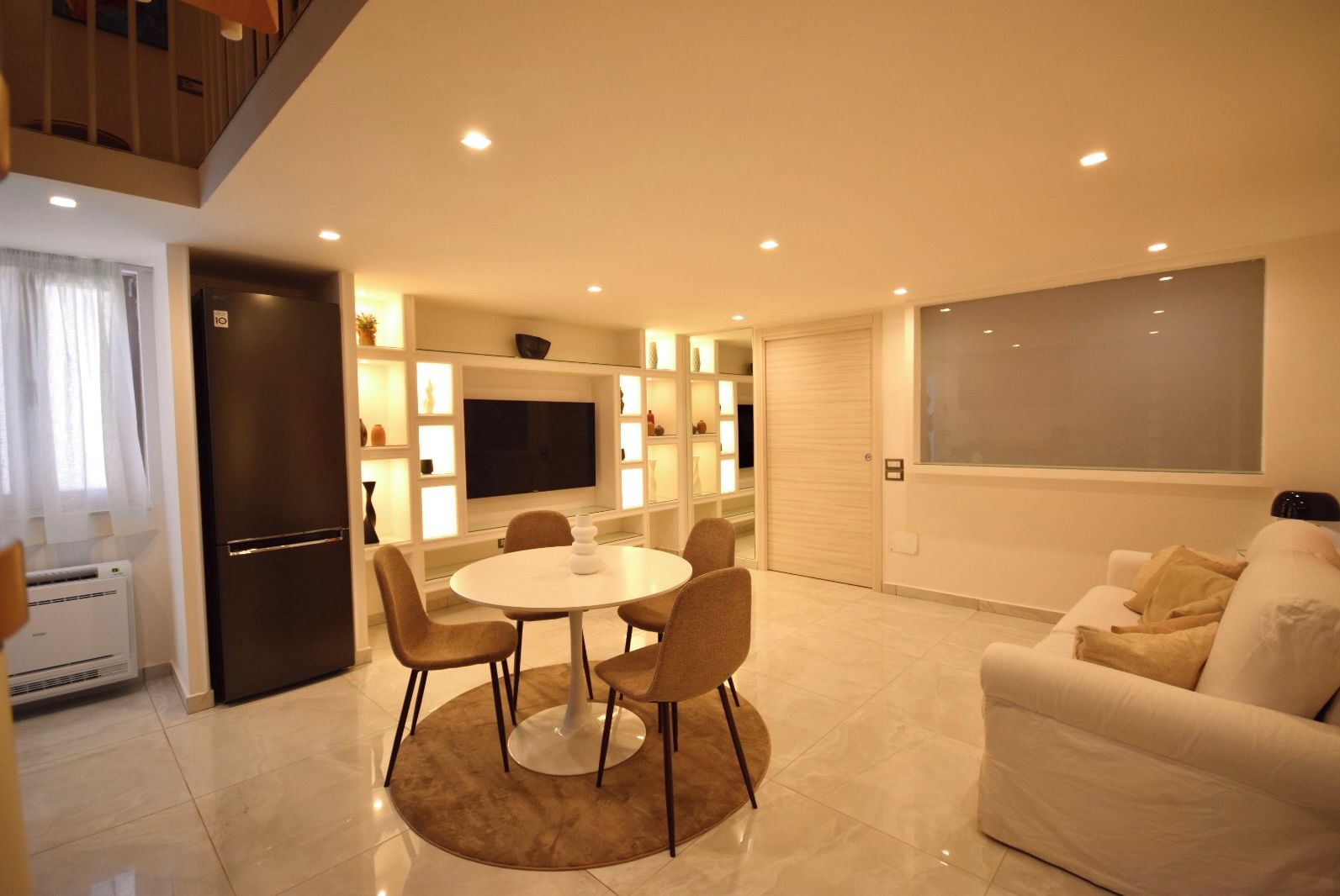 Un salon moderne avec un canapé blanc, une table à manger avec des chaises, un téléviseur à écran plat fixé au mur et un coin cuisine doté d’un éclairage intégré.