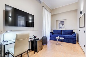 A modern living room with a flat-screen TV, a desk with a chair, a blue sofa, and a small table. - Muzio Private Suites by Premium Suites Collection (ROME)