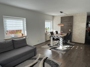 Un espai de vida modern amb un sofà gris, una taula de dinar amb cadires i una zona de cuina.