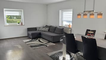 Ein modernes Wohnzimmer mit einem grauen Ecksofa, einem Esstisch mit Stühlen und einer Küchenzeile mit Fenster.