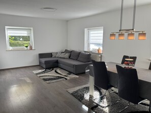 Ein modernes Wohnzimmer mit einem grauen Ecksofa, einem Esstisch mit Stühlen und einer Küchenzeile mit Fenster.