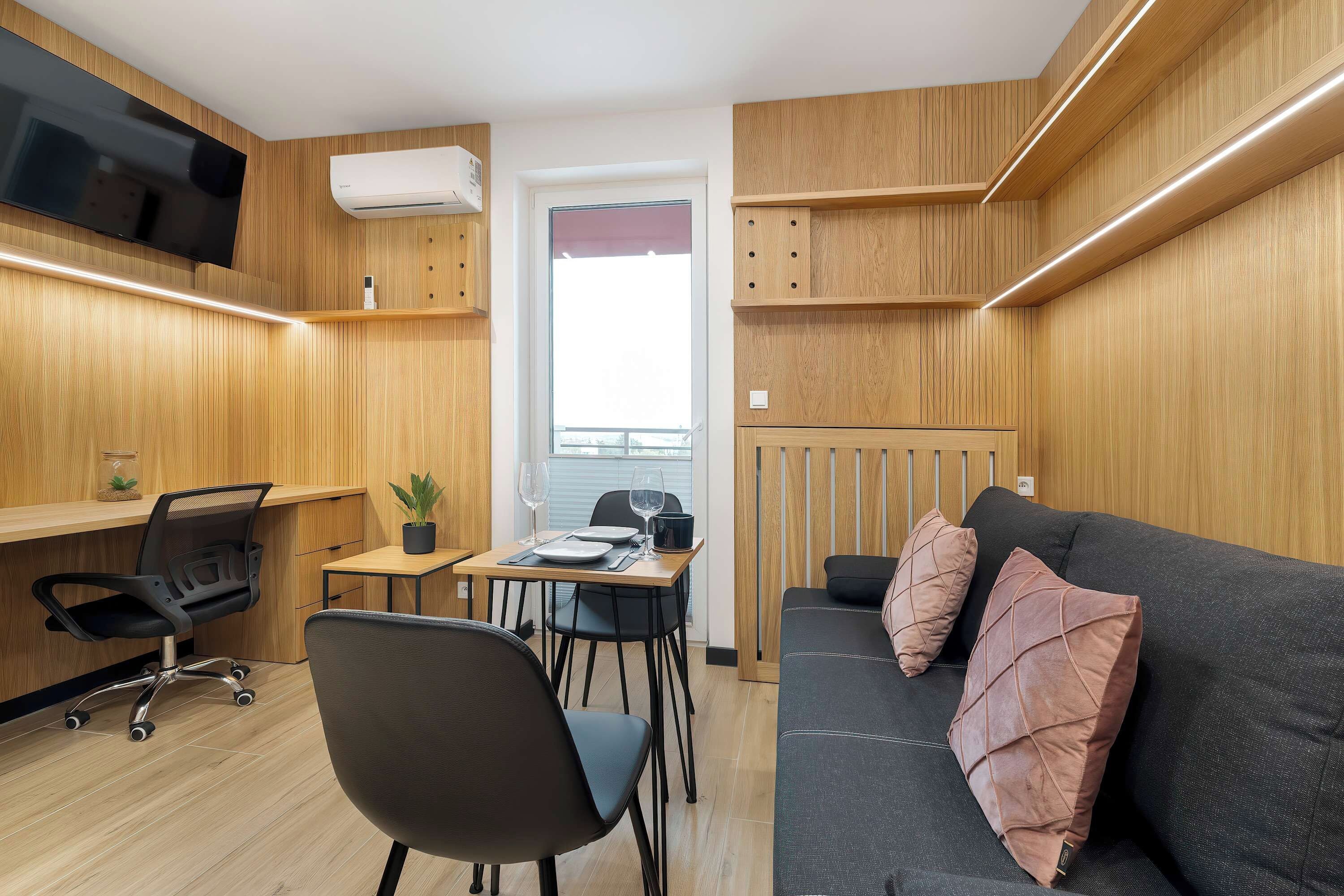 A compact room with a wooden interior, a sofa, a desk with a chair, a small table with a potted plant, and a television mounted on the wall.