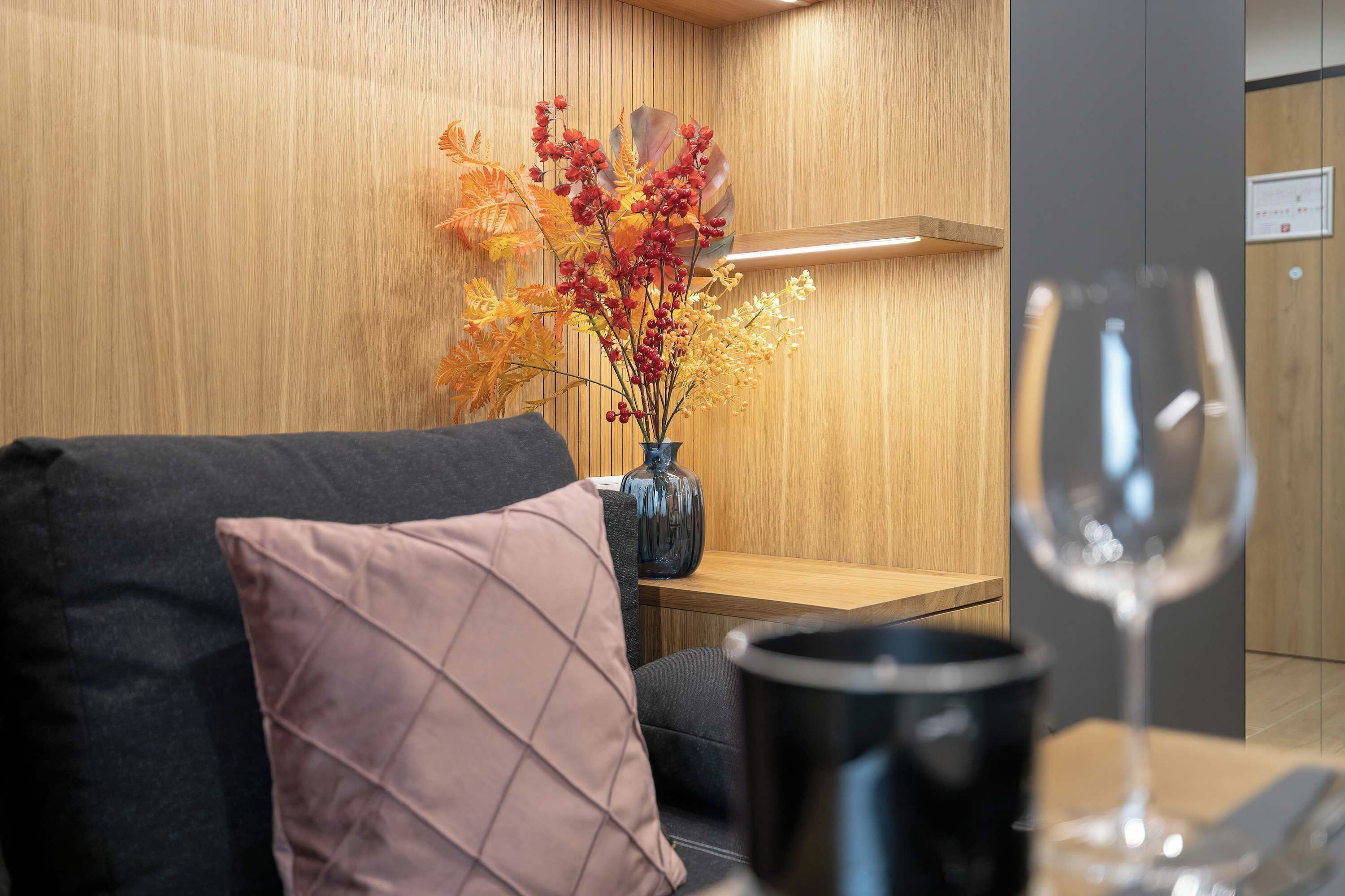 A modern interior with a wooden wall, a black sofa, a vase with dried flowers, and a glass of water on a table.