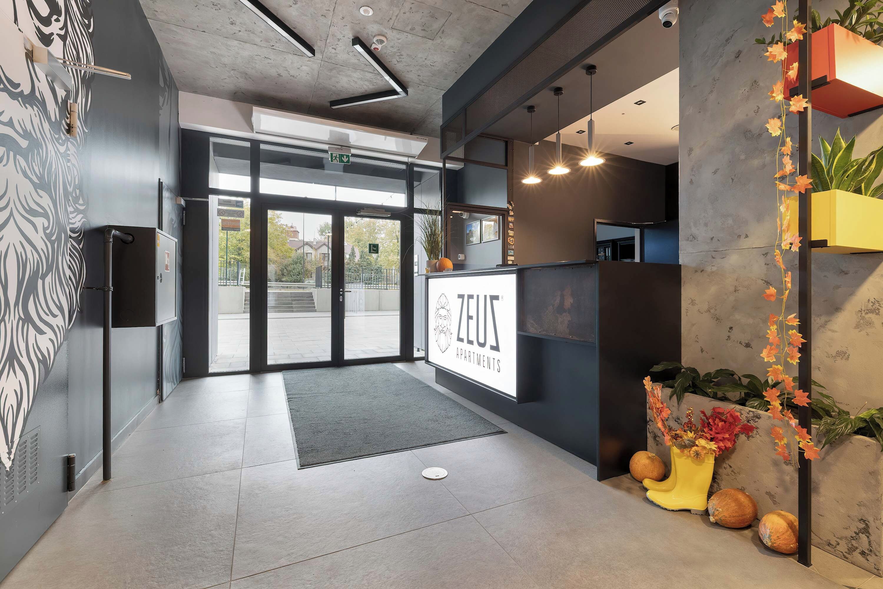 A reception area with a sign for 'ZEUS APARTMENTS,' a glass door entrance, a reception desk, and a decorative plant arrangement.