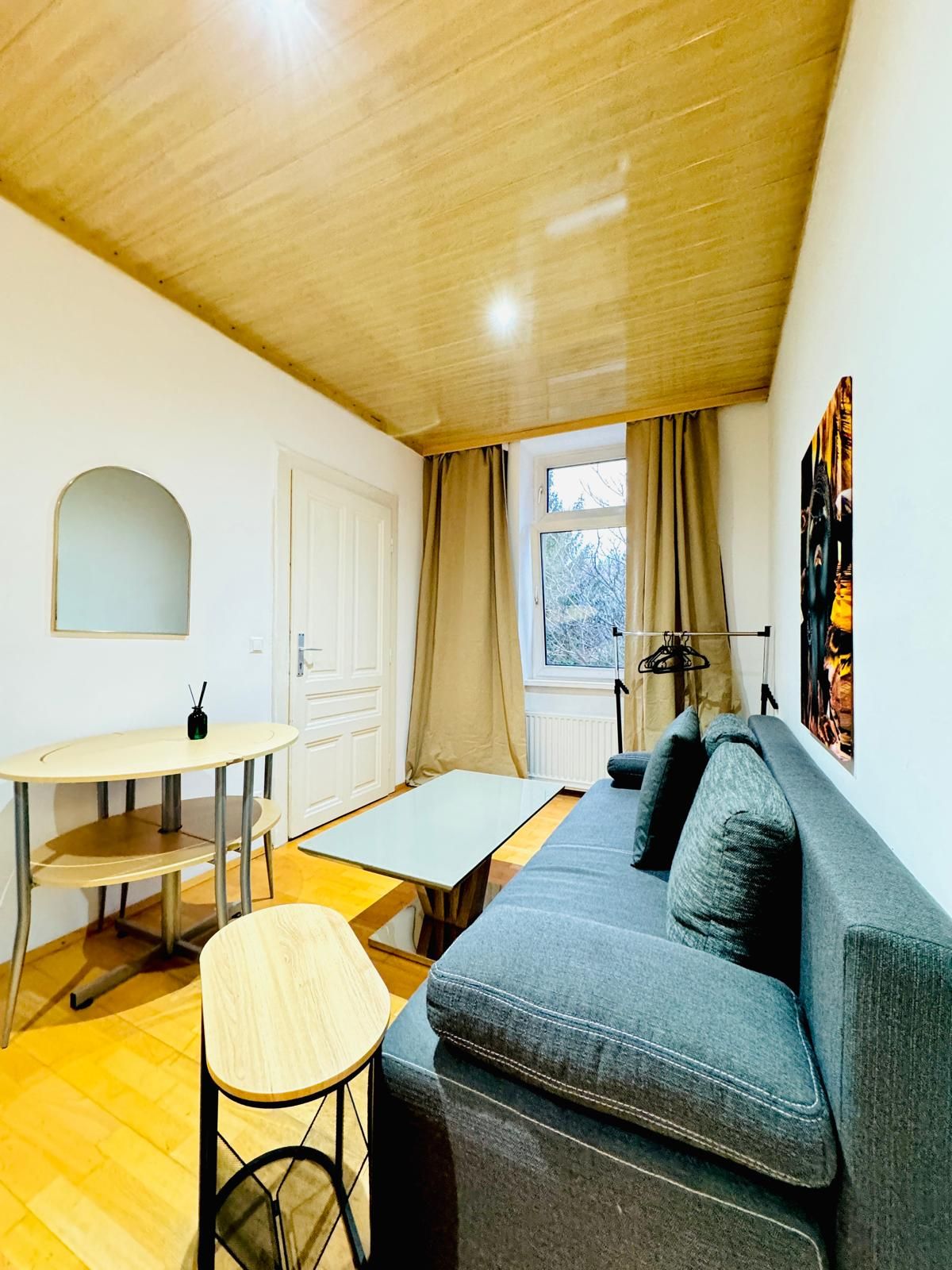 A compact living space with a wooden ceiling, a grey sofa, a small round table, and a window with curtains.