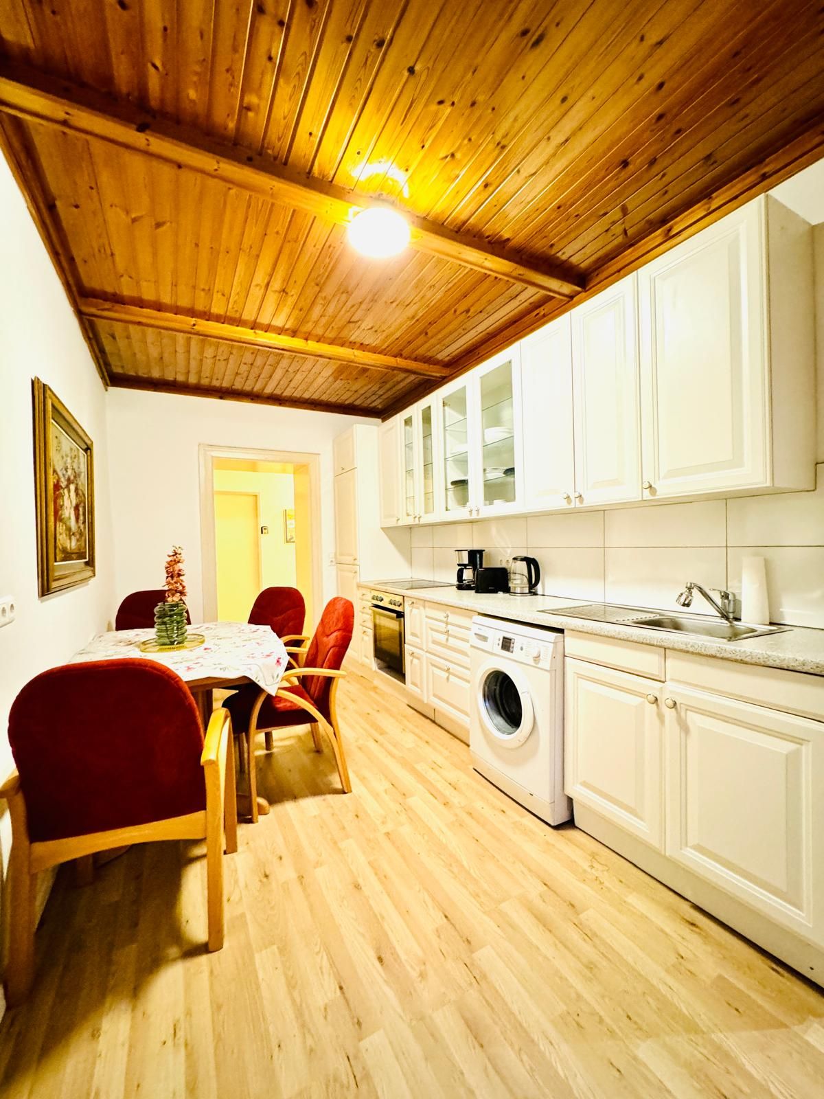 Een compacte keuken met houten balken in het plafond, witte kasten, een wasmachine en een eethoek met rode stoelen.