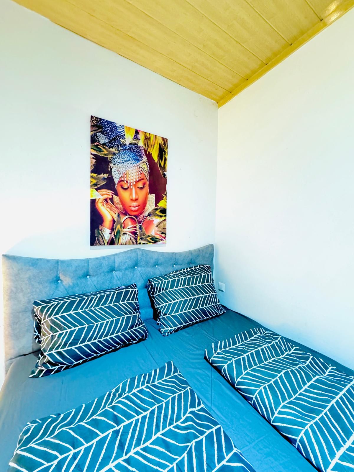 Een bed met een blauw hoofdeinde en gestreepte kussens, een muur met kleurrijke kunst, en een houten plafond.