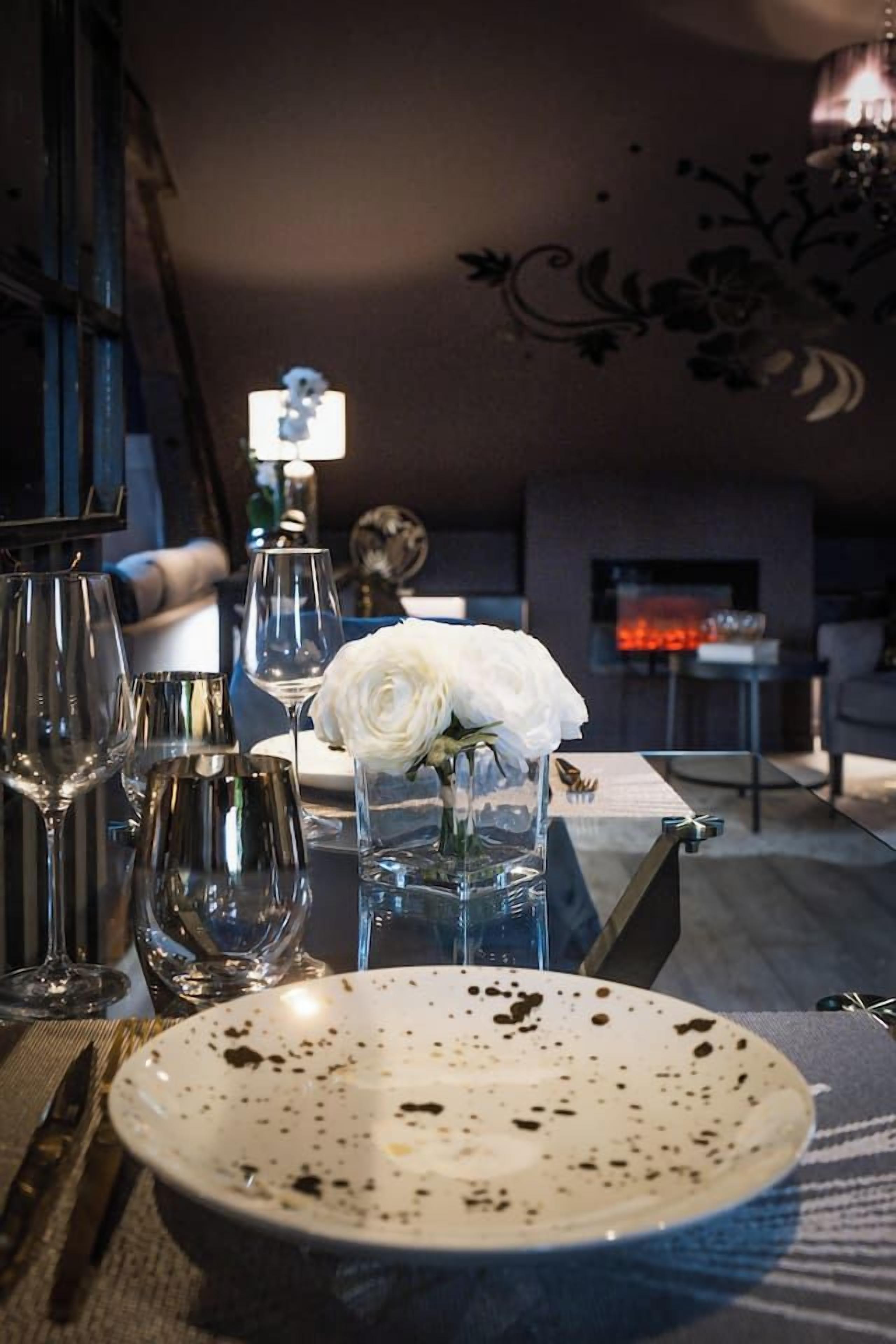 Une table à manger avec une assiette blanche, des verres à vin et un vase contenant une fleur blanche.