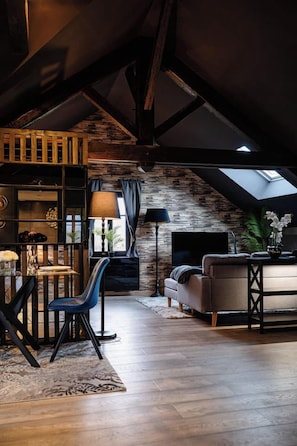 Een loft-achtige woonruimte met zichtbare balken, een stenen muur, een bank, een televisie en een eethoek met een tafel en stoelen.