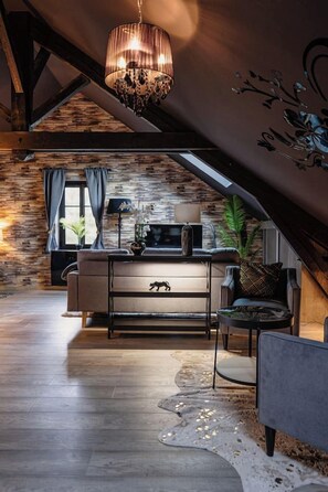 A cozy loft-style room with exposed wooden beams, a stone wall, a modern sofa, a chandelier, and a large window with curtains.