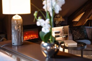 Een gezellige woonkamer met een open haard, een lamp, een vaas met bloemen en een bank met decoratieve kussens.