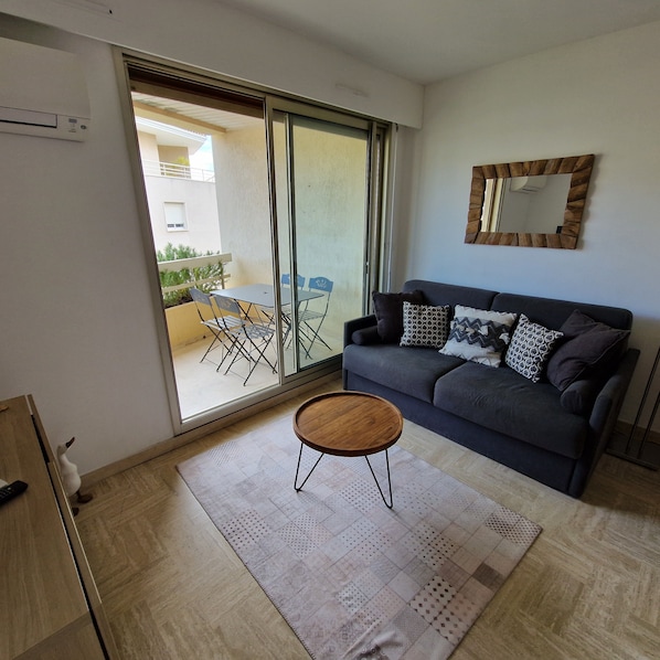 A living room with a sofa, a coffee table, a sliding glass door leading to an outdoor area with chairs and a table. - Charming Air-Conditioned Studio with Balcony, Steps from the Beach (Cavalaire-sur-Mer)