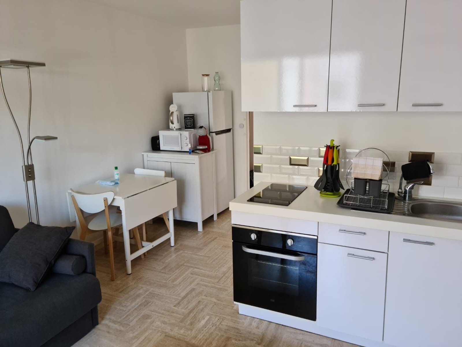 Cocina compacta con muebles blancos, un microondas, un hervidor y un lavamanos. Hay una zona de comedor con una silla y una mesa, y un sofá en primer plano.