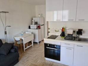 A compact kitchen with white cabinetry, a microwave, a kettle, and a sink. There's a dining area with a chair and a table, and a sofa in the foreground. - Charming Air-Conditioned Studio with Balcony, Steps from the Beach (Cavalaire-sur-Mer)