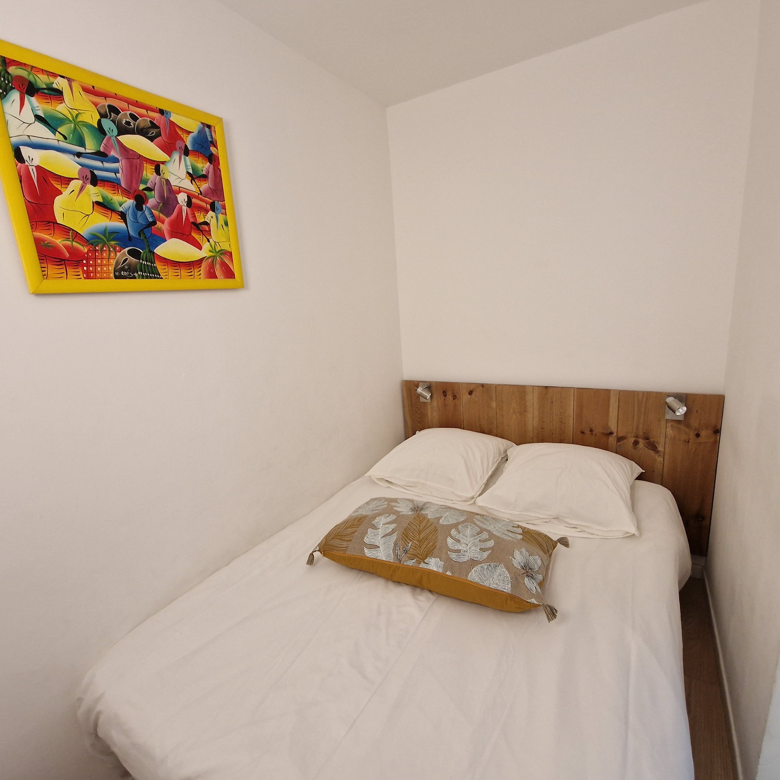 Una cama individual con ropa de cama blanca y una almohada decorativa, un cabecero de madera y un cuadro colorido en la pared.