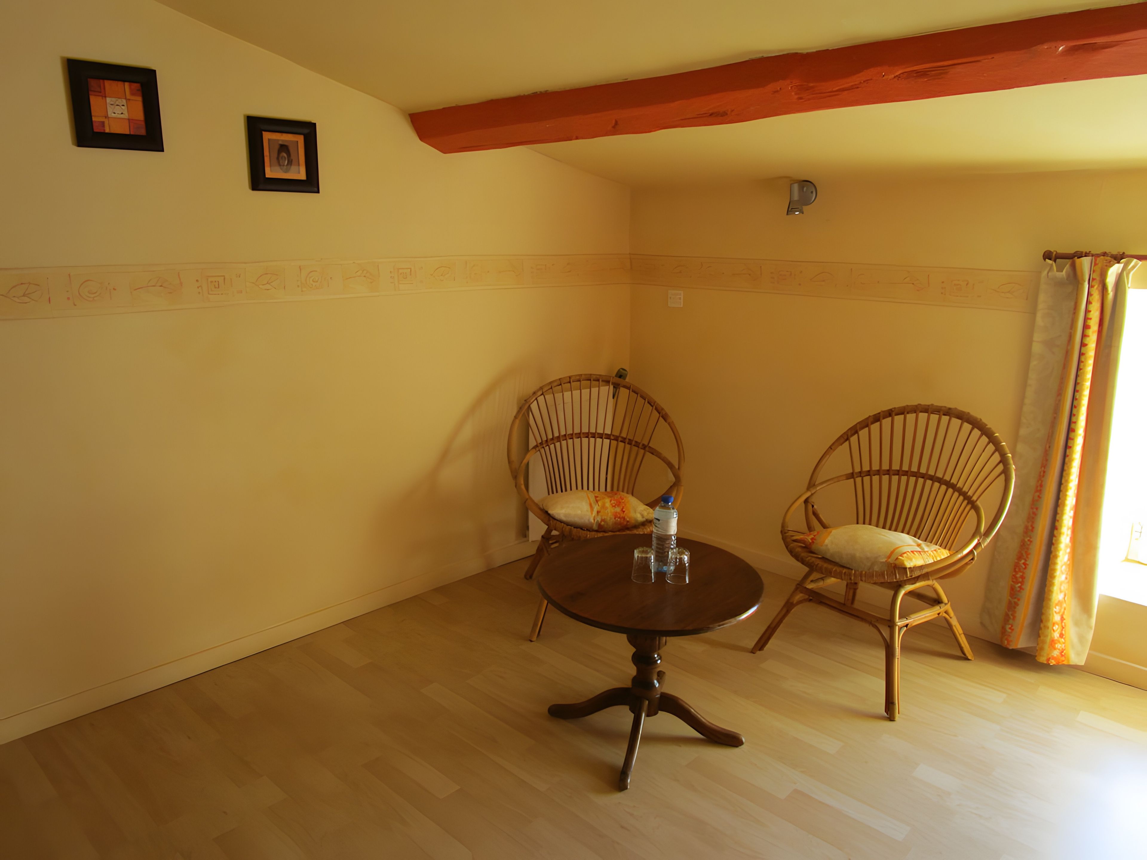 Une pièce avec deux fauteuils en osier, une table ronde, un plancher en bois et une fenêtre avec des rideaux.