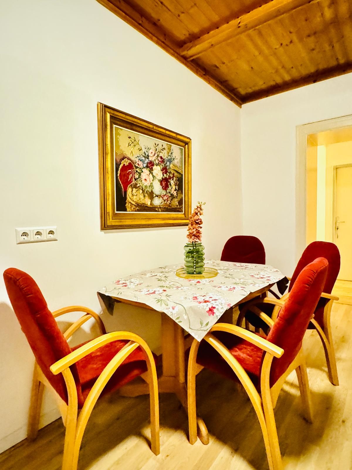 A dining area with a wooden table and four red upholstered chairs. A painting hangs above the table, and a vase with flowers is on the table.