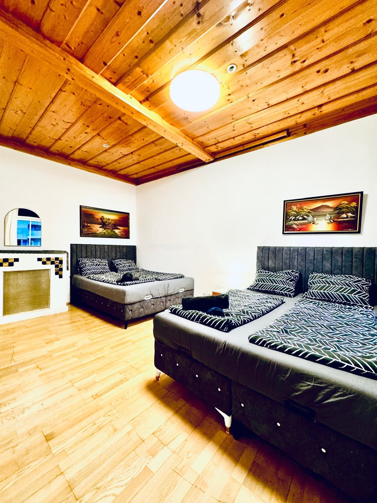 A room with a wooden ceiling, two beds with patterned bedding, a small window, and two wall-mounted paintings.
