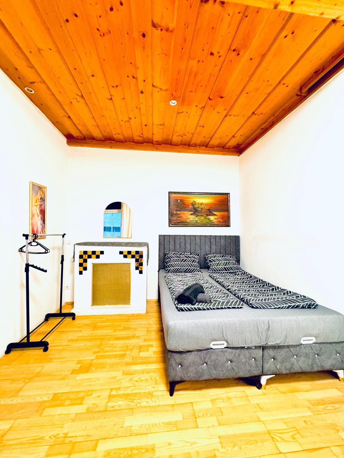 Een slaapkamer met een houten plafond, een bed met beddengoed met een patroon, een klein tafeltje met een ruitjesdessin en een wandradiator.