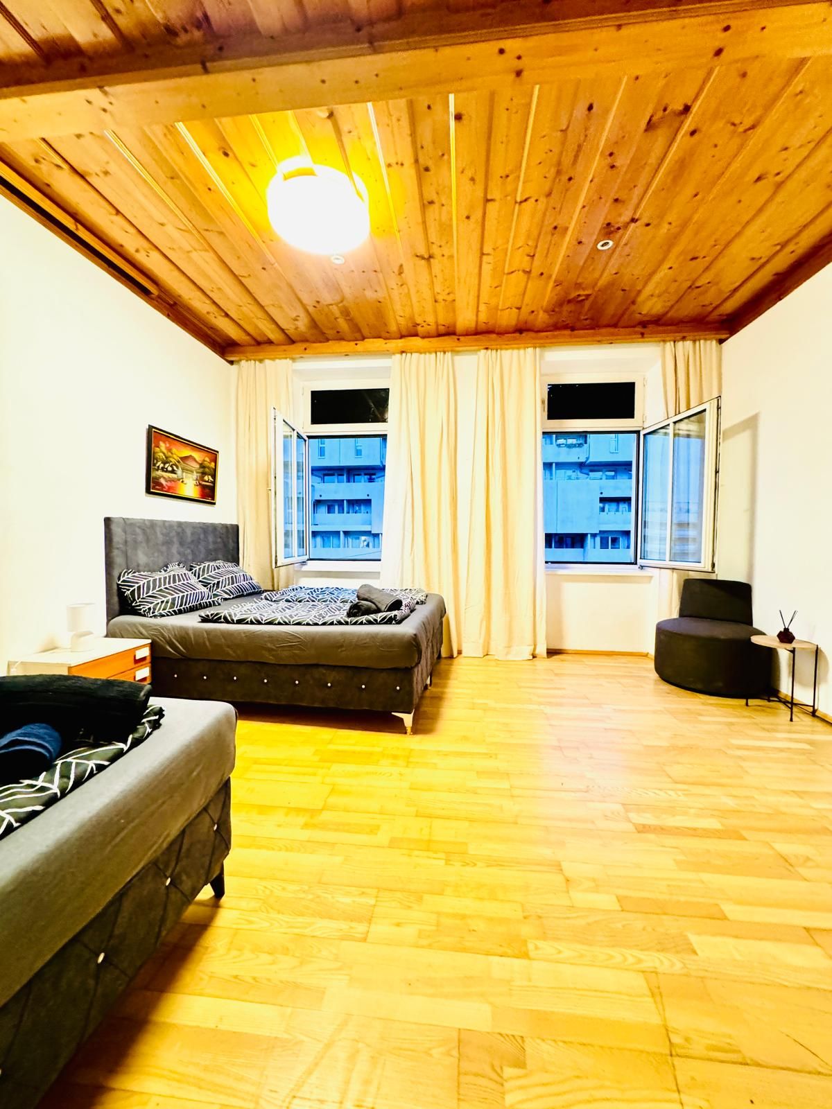 Een gezellige kamer met een bed, een bank en een stoel. Twee ramen met gordijnen, een plafondlamp en een schilderij aan de muur.