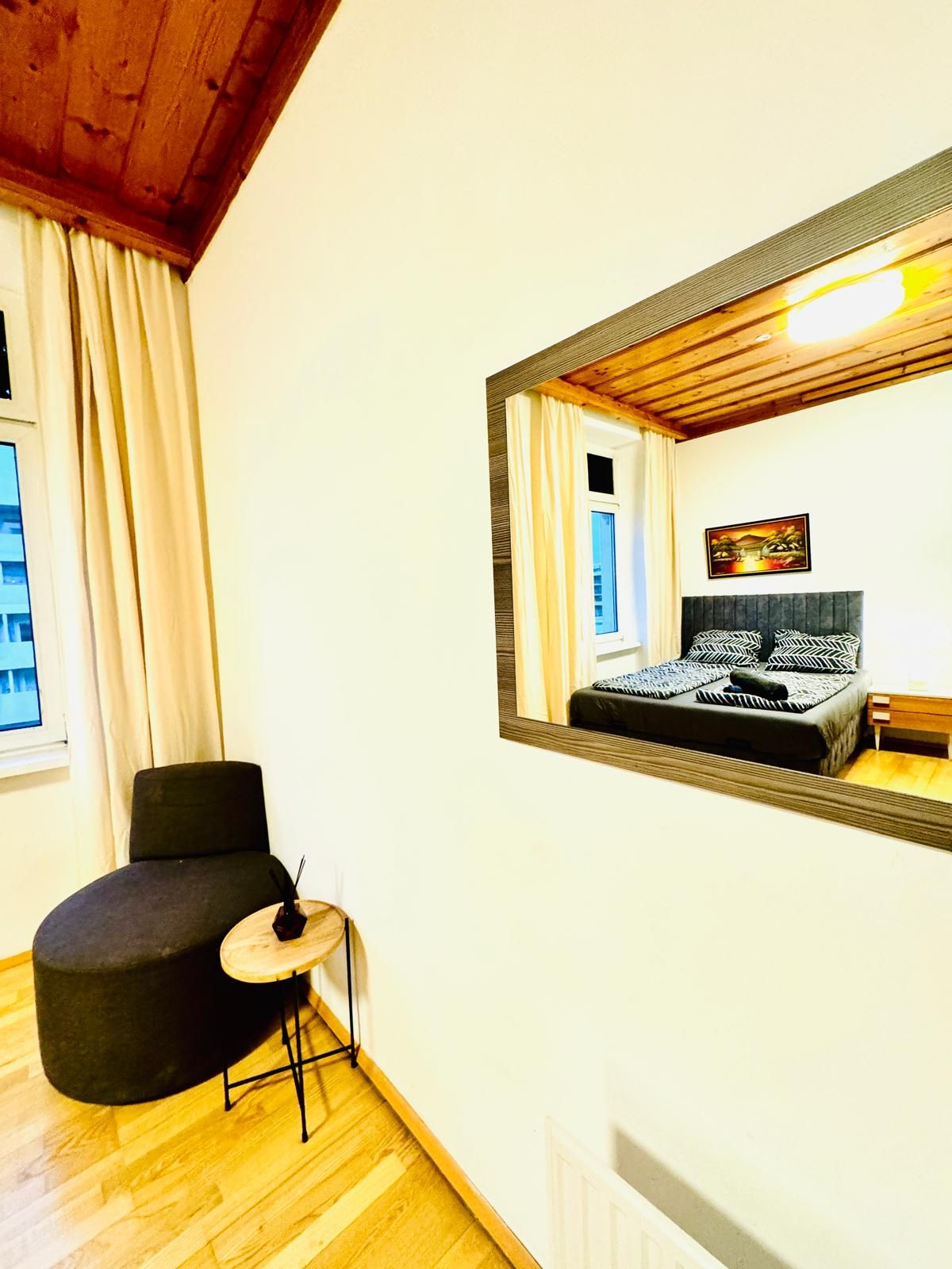 A bedroom with a large mirror, a bed with patterned bedding, a chair, a small table, and a window with curtains.