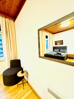 A bedroom with a large mirror, a bed with patterned bedding, a chair, a small table, and a window with curtains. - Vienna Basecamp Private Room (Wien)