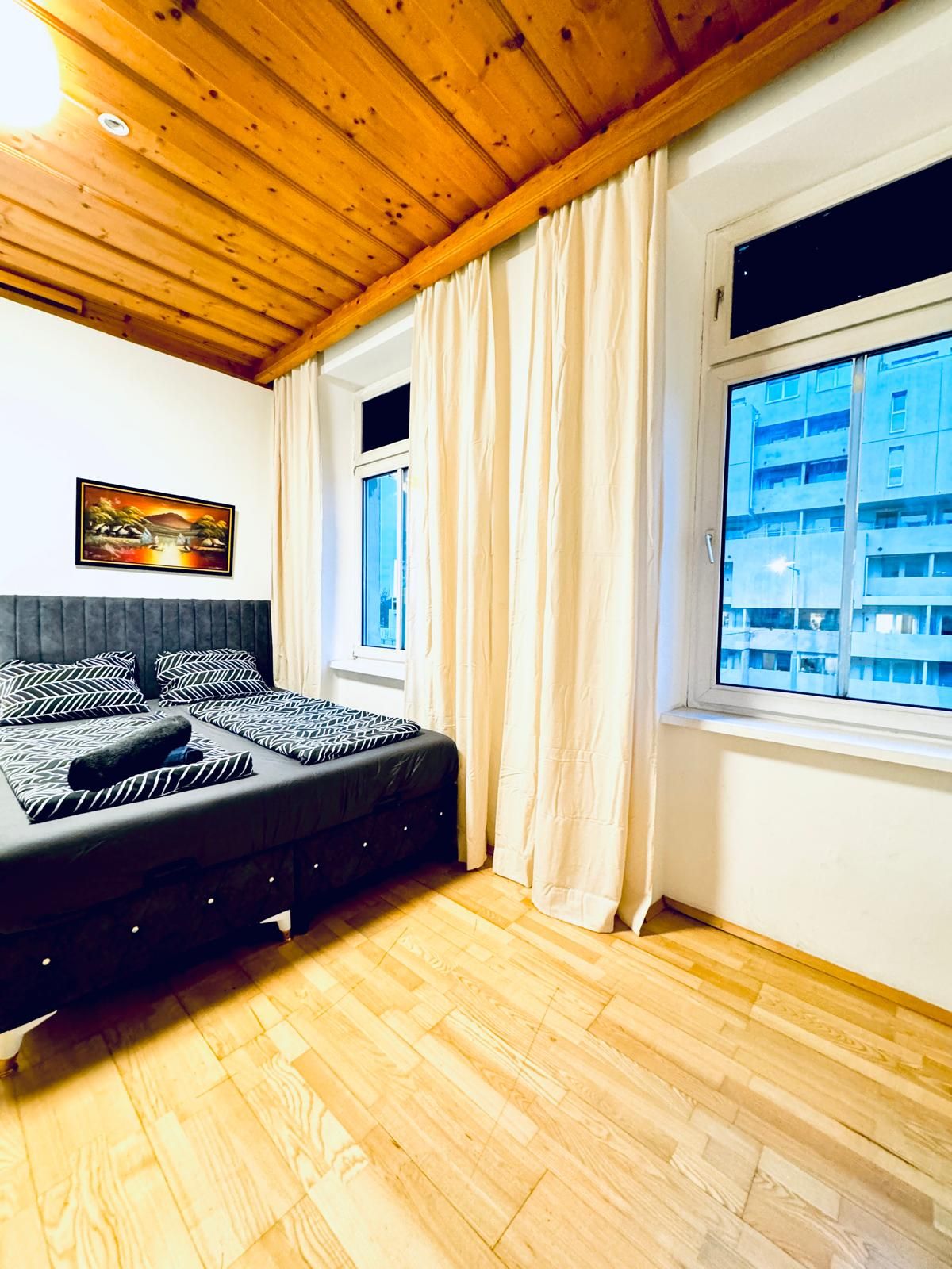 A bedroom with a bed, wooden floor, a window with curtains, and a painting on the wall.