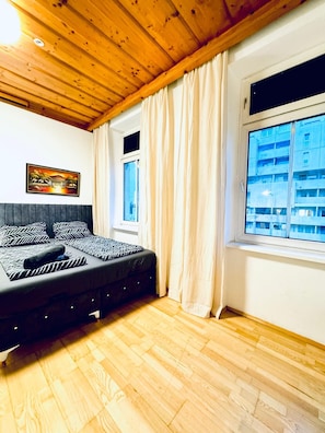 Une chambre à coucher avec un lit, un plancher en bois, une fenêtre avec des rideaux et un tableau accroché au mur.