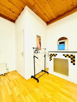 A room with a wooden ceiling, a white door, a radiator, a black metal rack, a painting, and a checkered fireplace. - Vienna Basecamp Private Room (Wien)