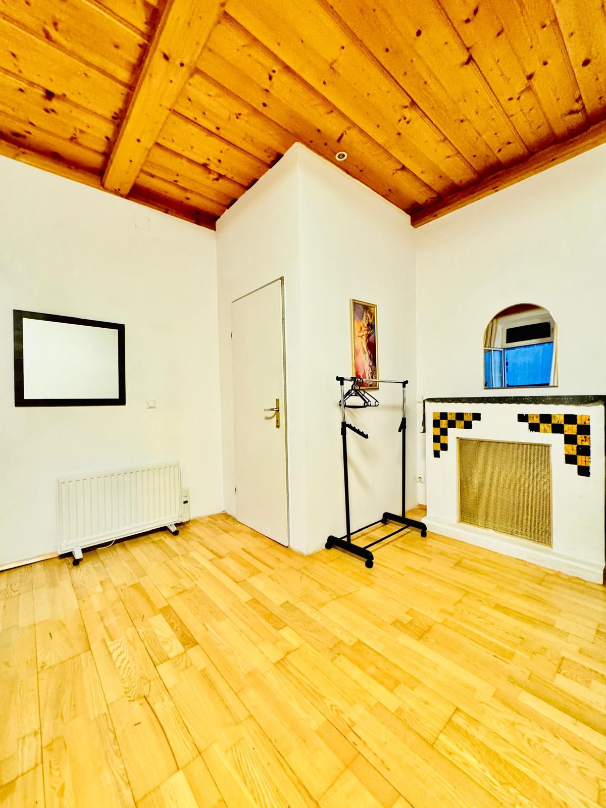 Een kamer met houten vloer, een witte radiator, een deur, een kapstok, een lijst met een afbeelding en een haard met een geruit patroon.