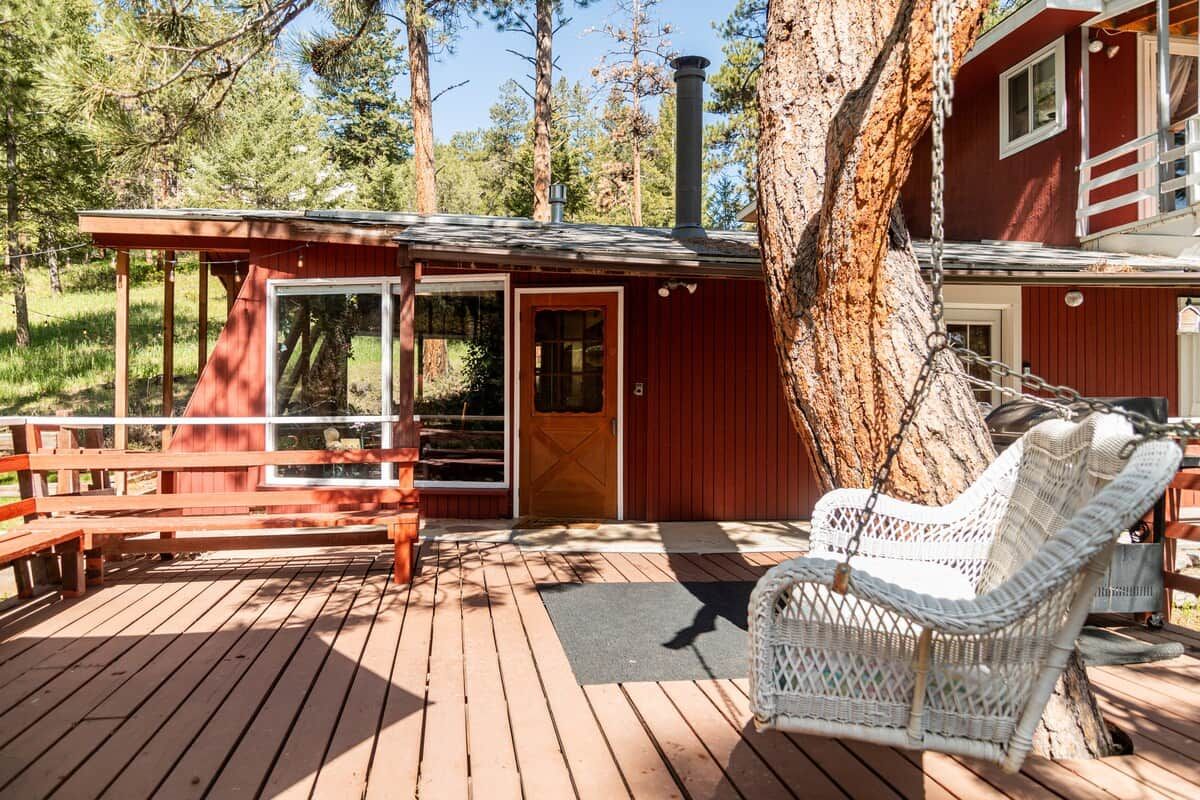 A wooden deck with a wicker chair and a bench, a red building with a chimney, and a large tree.