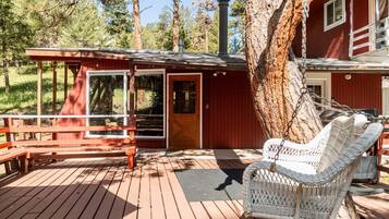 A wooden deck with a wicker chair and a bench, a red building with a chimney, and a large tree.