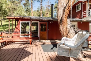 A wooden deck with a wicker chair and a bench, a red building with a chimney, and a large tree. - Aspen Song Mountain Retreat (Jefferson County)