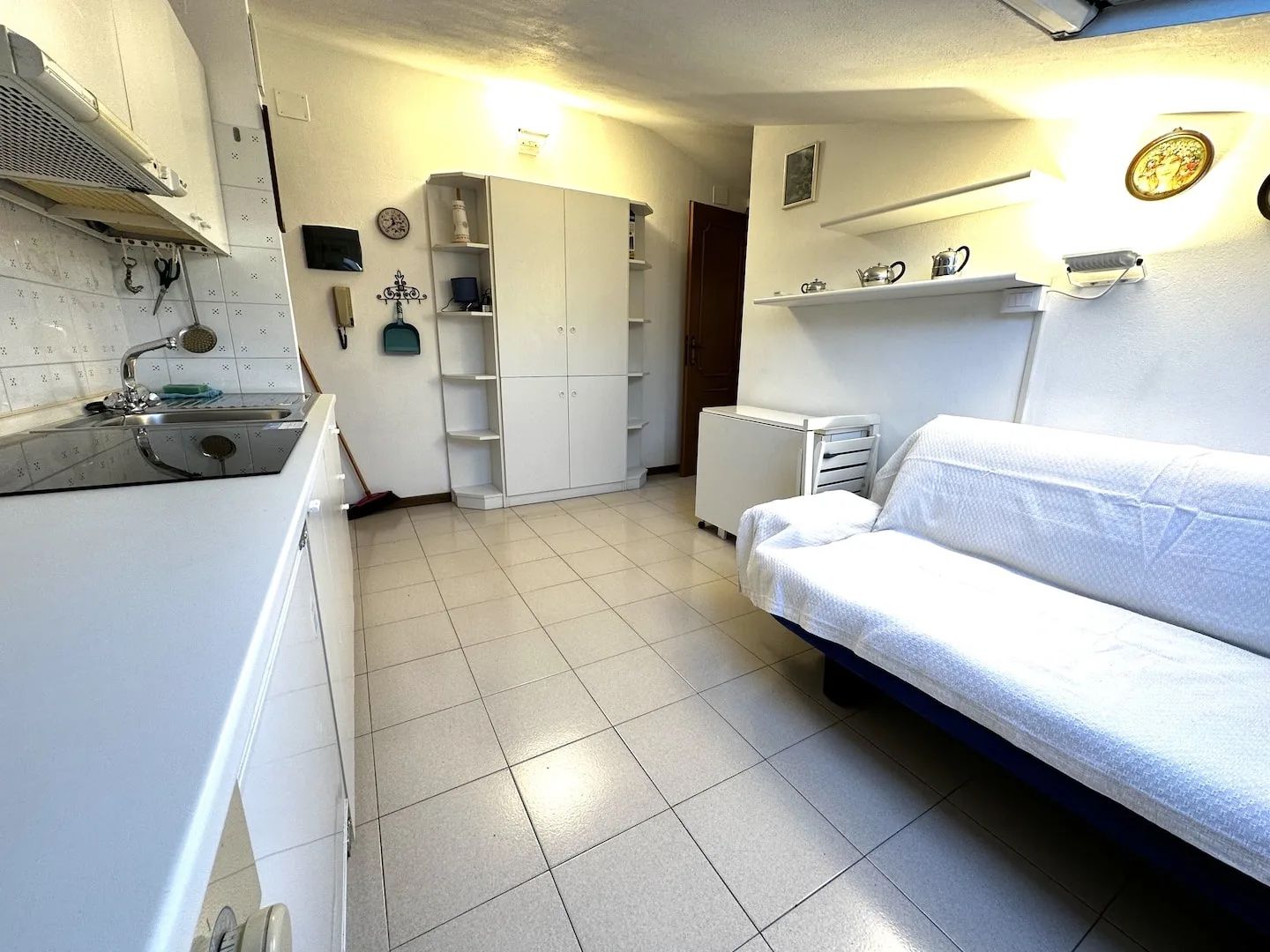 A compact kitchen with a sink, cabinets, and a small countertop. There is a white sofa and a radiator in the room.