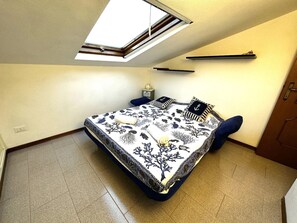 Une chambre avec une lucarne, un lit recouvert d’une couvre-lit sur le thème de la marine, une table de chevet et une petite table de nuit.