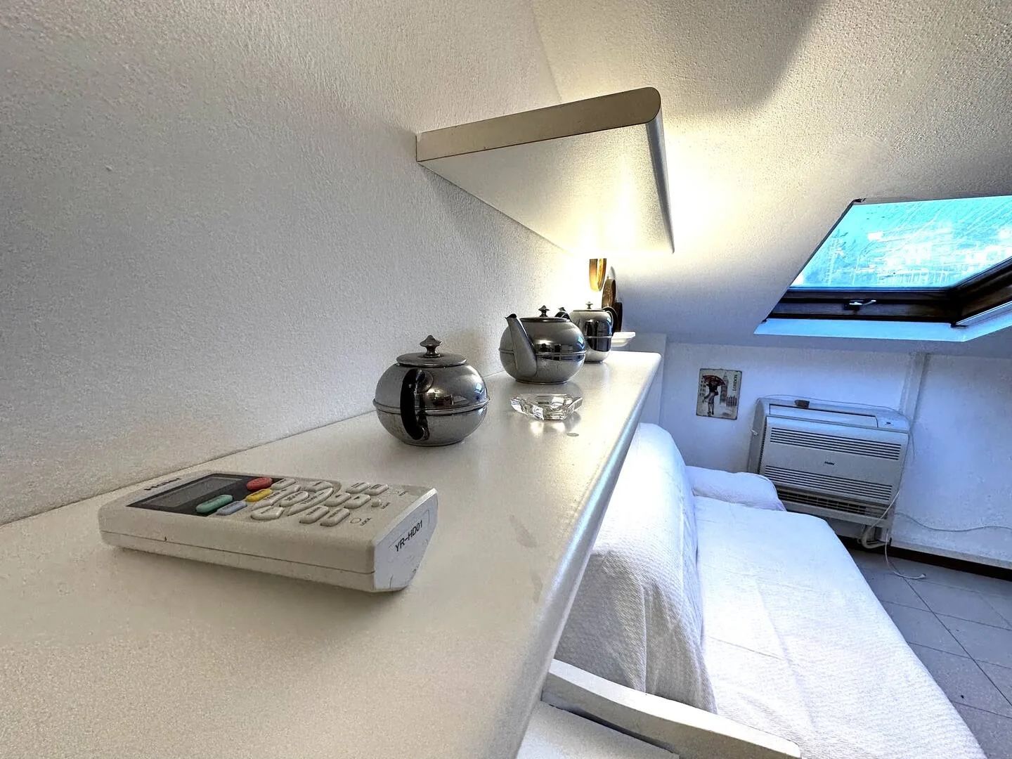 A hotel room with a bed, a bedside table with a digital clock, a small table with a teapot and cups, and a window with a view.