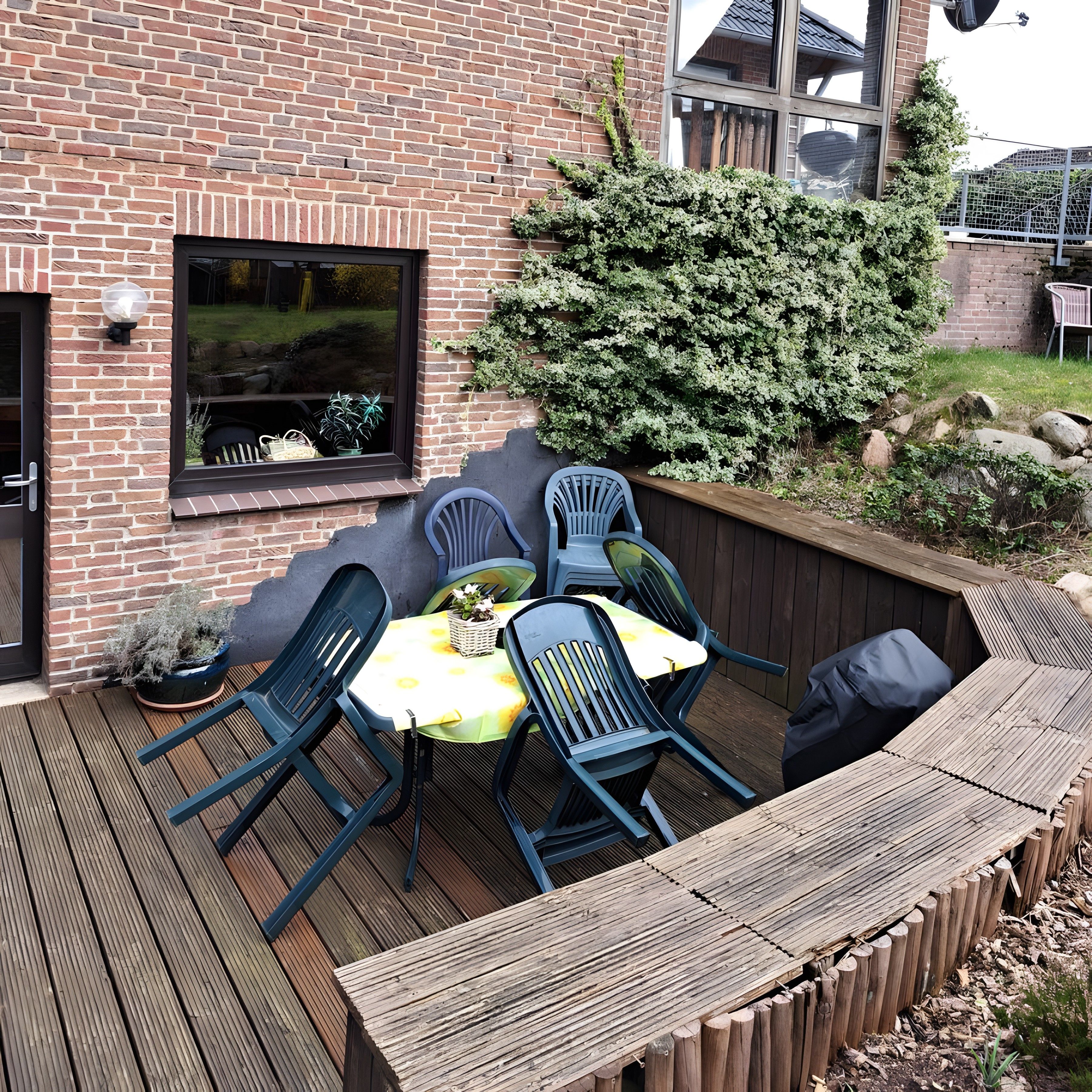 Eine Holzterrasse mit Tisch und Stühlen, eine Backsteinmauer und ein Fenster.