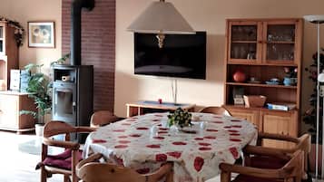 Een eethoek met een tafel voor vier personen, omringd door houten stoelen, een aan de muur bevestigde televisie en een open haard met bakstenen.