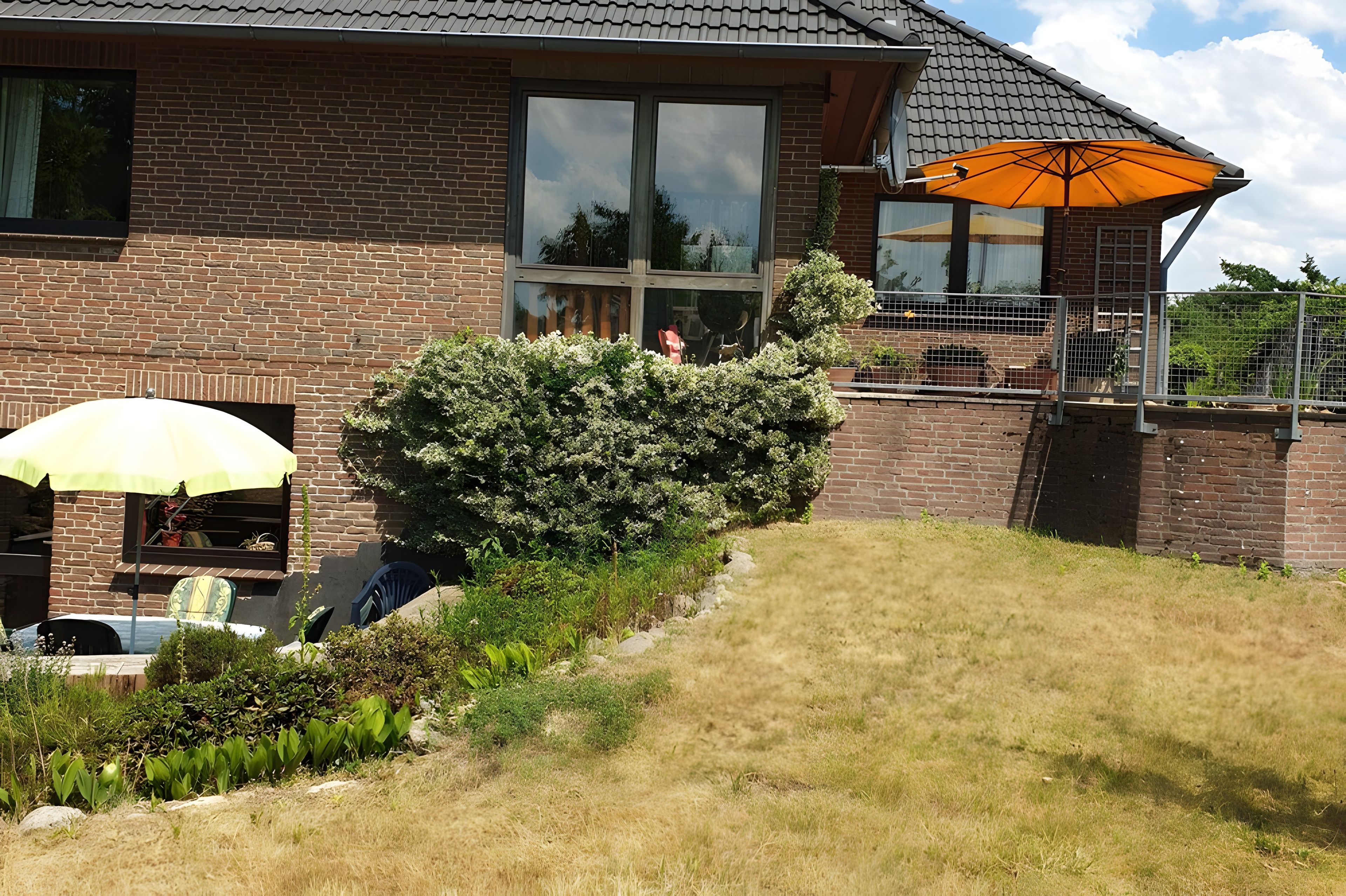 Ein Ziegelhaus mit großem Fenster, einer Terrasse mit gelber Sonnenschirm und einem gepflegten Garten.