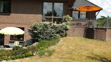 Een bakstenen huis met een groot raam, een terras met een gele parasol en een verzorgde tuin.