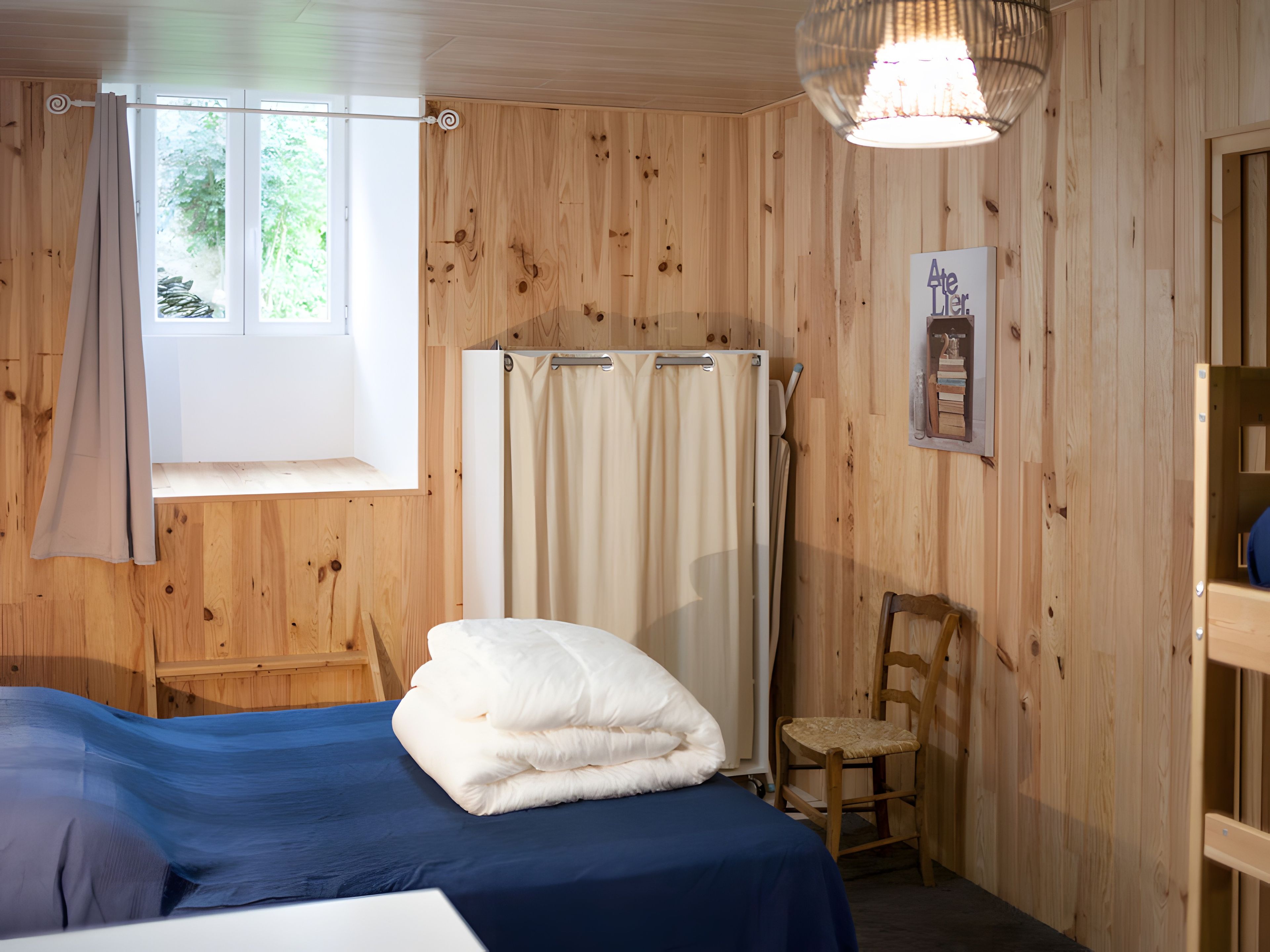 Ein Zimmer mit Holzwänden, einem Bett mit blauer Bettdecke, einem weißen Kopfkissen, einem Holzstuhl, einem Duschvorhang und einer hängenden Leuchte.