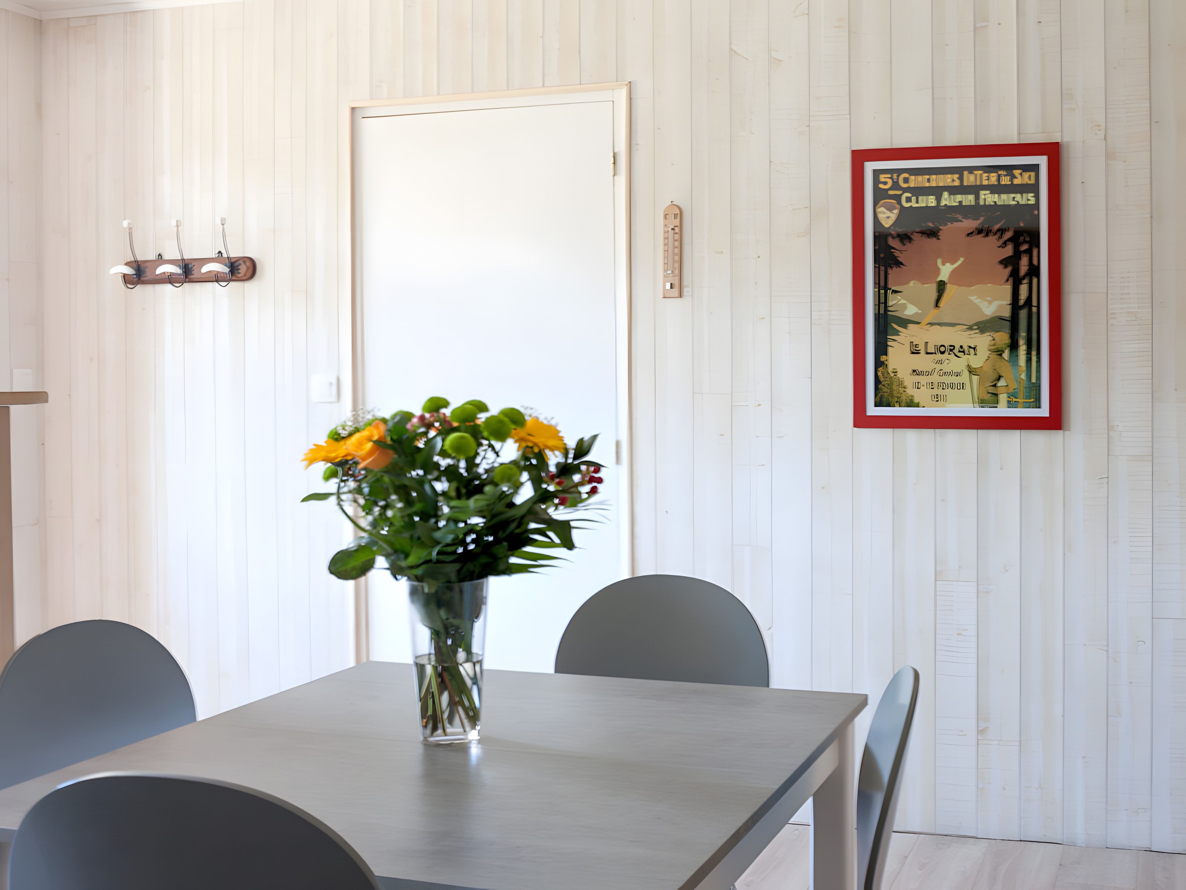 Uma sala de jantar com uma mesa, cadeiras, um vaso de flores, uma porta e um cartaz emoldurado na parede.