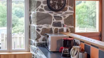 Une cuisine avec un mur en pierre, une horloge, une machine à café et une vue sur la verdure à l’extérieur.