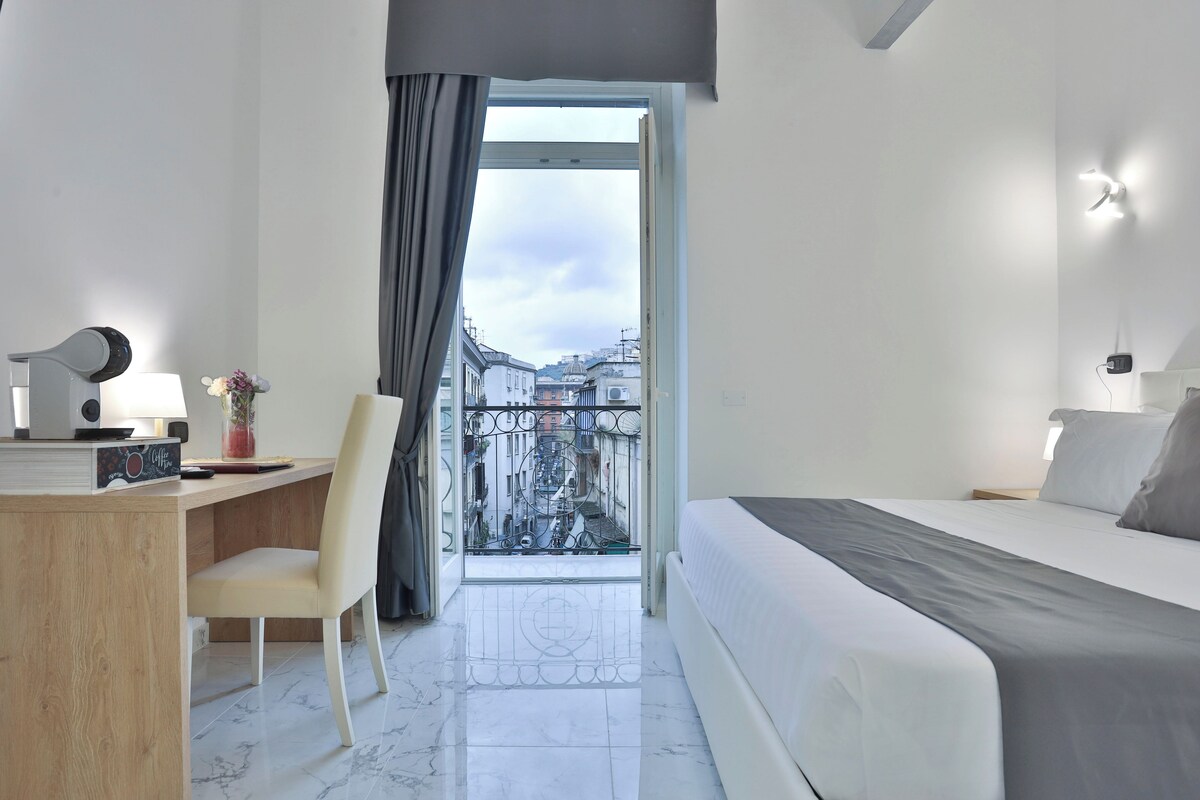 A hotel room with a bed, a desk, a chair, a window with a view of buildings, and a curtain.
