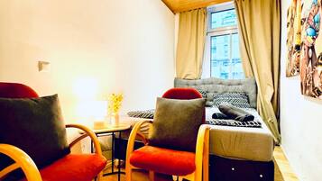 Habitació petita i acollidora amb sostre de fusta, un llit amb roba de llit estampada, dues cadires amb coixins, una taula petita amb un ram i una finestra amb cortines.