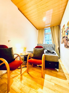 Ein kleiner, gemütlicher Raum mit Holzbalkendecke, einem Bett mit gemusterten Textilien, zwei Sesseln mit Kissen, einem kleinen Tisch mit einer Vase und einem Fenster mit Vorhängen.